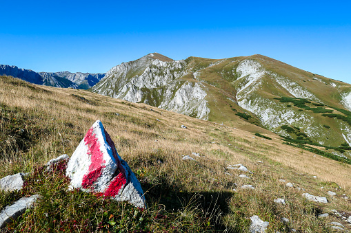 A red-white-red path mark along the way to Hohe Weichsel, Austrian Alps. Sharp peaks around. There is a vast pasture on top of a mountain, slowly turning golden. Clear, blue sky above. Autumn vibe