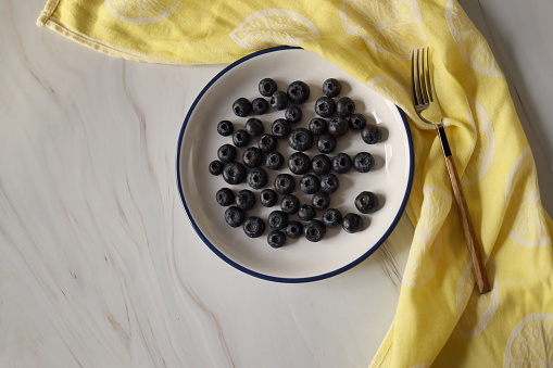 Istanbul, Turkey-March 14, 2021: Blueberries on white ceramic plate on white-gray marble floor, Next to the plate is a yellow linen napkin and fork, The edge of the plate has a dark blue stripe, Full Frame, Still life, Studio shot, Flat lay. Shot with Canon EOS R5.