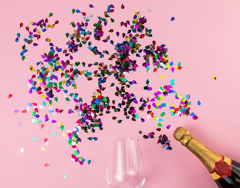 Wine glass, bunch of confetti and champagne bottle on pink background. Flat lay concept.