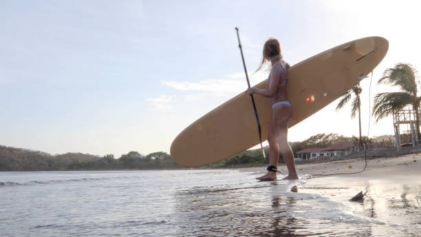 istock Young woman walks down beach with paddle board in the morning 1308142321