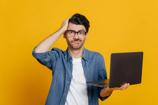 Frustrated displeased unshaven caucasian guy in glasses and in denim shirt, holds a laptop, looks confusedly at the camera, holding his head, without mood, stands on an isolated orange background