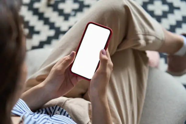 Photo of Young woman or teen girl hands holding mobile smart phone with mockup white blank display, empty screen at home. Shopping, delivery apps, social media applications ads, over shoulder close up view.