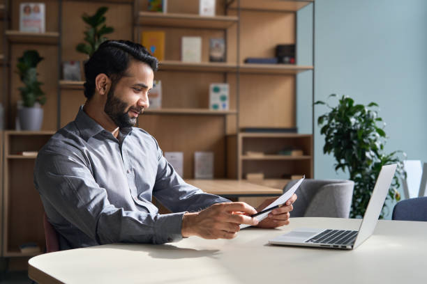 istock Smiling indian business man employee working on laptop looking at document at home office. Male ethnic professional teacher reading paper, employer holding cv, lawyer checking contract sits at table. 1308168032