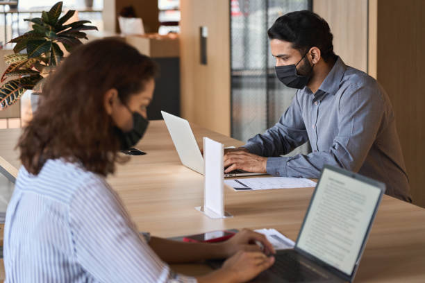 istock Indian business man student wearing face mask working on laptop safe distancing. Diverse people in facemasks using computers sitting at table with social distance sign in office coworking or cafeteria 1308168037