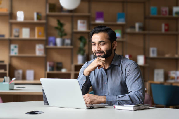 lächelnd bärtige indische geschäftsmann arbeiten auf laptop in modernen büro-lobby-bereich. junge indische student mit computer-remote-studium, beobachten online-webinar, virtuelles training auf video-anruf-meeting. - people reading stock-fotos und bilder