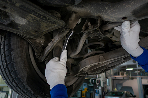 At a repair shop, a car mechanic tightens the suspension of an elevated vehicle with a spanner.