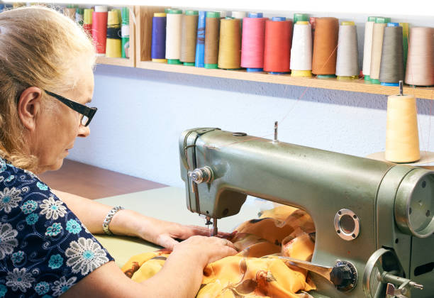 zdjęcie boczne starszej kobiety w okularach szyjących żółtą wzorzyste tkaniny z przemysłową maszyną do szycia oświetloną od góry półką kolorowych nici - costurera zdjęcia i obrazy z banku zdjęć