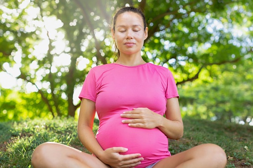 A happy pregnant woman sitting on the grass at the park with her eyes closed relaxing and meditating for health and stress.