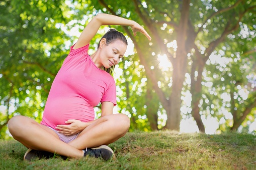 A pregnant mom sitting on the grass stretching and staying fit, practicing yoga.