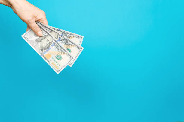 woman's hand holding out one hundred dollar bills from above. blue background, copy space - over 100 number 100 senior adult women imagens e fotografias de stock