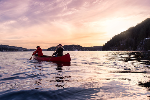 Couple friends on a wooden canoe are paddling in water. Dramatic Sunset Sky Art Render. Taken in Indian Arm, near Deep Cove, North Vancouver, British Columbia, Canada. Concept: Adventure, Explore