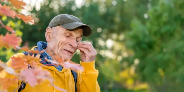Old man in cap kills mosquito and touches bite place on face standing in forest.