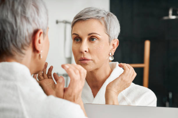 femme aîné avec la crème hydratante sur son visage regardant le miroir dans la salle de bains - miroir photos et images de collection