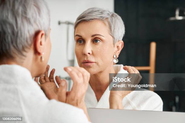 Senior Woman With Moisturizer On Her Face Looking At Mirror In Bathroom Stock Photo - Download Image Now