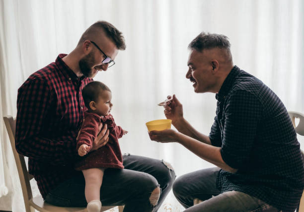 männliche homosexuell paar mit adoptierten baby mädchen zu hause - zwei schöne väter füttern das baby mädchen auf küche - männliche babysitter - lgbt familie zu hause - diversity-konzept - homosexual stock-fotos und bilder