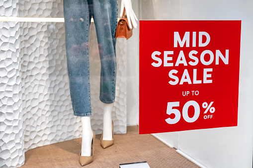 Close-up shot of red mid-season sale up to 50% stickers on retail store window with a background of the female mannequin on a nude stiletto and denim jeans, carrying a brown sling bag.