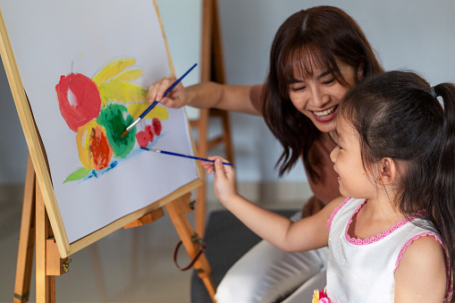 An Asian little girl painting with her mother using easel stand during art class at art studio.