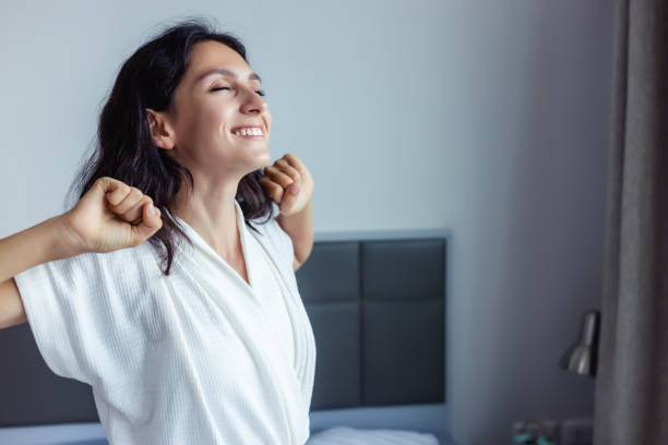 hermosa mujer despertando en su cama en el dormitorio joven hembra estirando y sonriendo encantado belleza dama despertar encontrarse buen fin de semana temprano por la mañana joven señora se sienten optimistas en casa con cara de sonrisa - levantarse fotografías e imágenes de stock