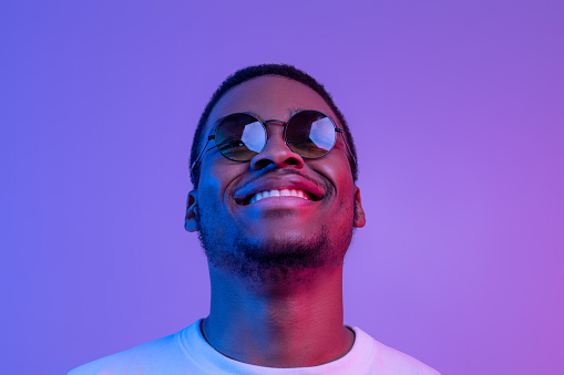 Closeup Shot Of Dreamy African American Man In Stylish Sunglasses Looking Up While Posing Under Neon Lighting, Smiling Black Millennial Guy Standing Over Purple Background, Free Space