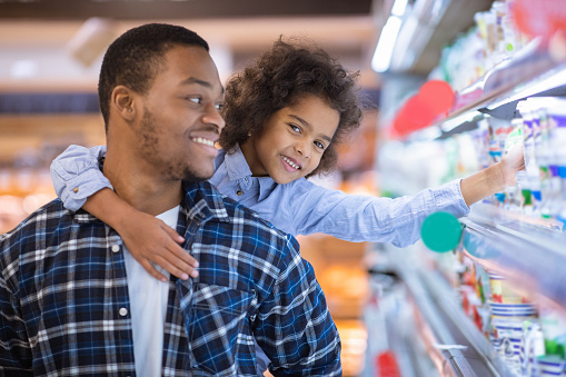 Young black father and his adorable daughter shopping for dairy products at supermarket, copy space. Cute girl with her daddy buying groceries at mall, selecting foodstuff together