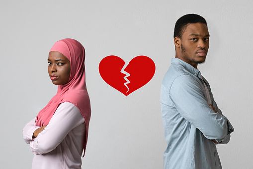 Disappointed black man and woman standing back to back with broken heart between them over grey studio background. Breakup, difficulties in relationships, marriage crisis concept