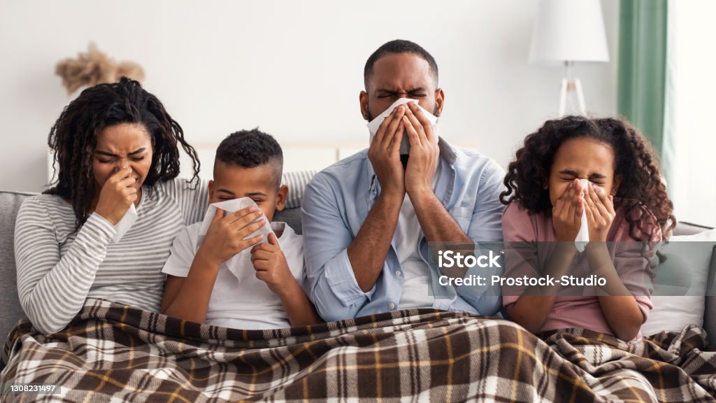 Sick black family blowing runny noses with napkins together Portrait of sick young black family of four people blowing runny noses while sitting together on the sofa with napkins and covered with blanket. African American parents and kids suffering from cold Allergy Stock Photo