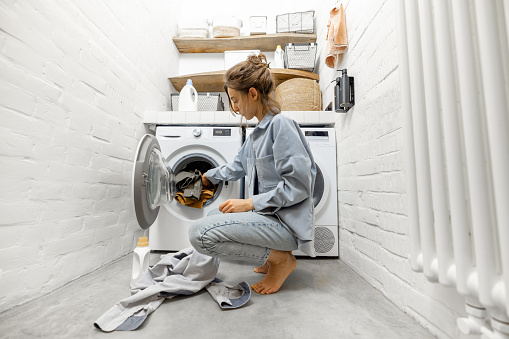 Young woman loads the washing machine with dirty color clothes at the laundry room at home, wide angle view