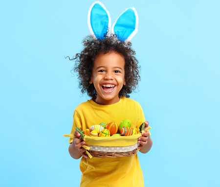 Adorable happy little African American boy with curly hair in casual clothes and bunny ears on head laughing while holding Easter basket