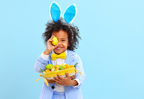 Cheerful little ethnic boy with Afro hair in Easter costume and bunny ears smiling while covering eye with colorful egg against blue background