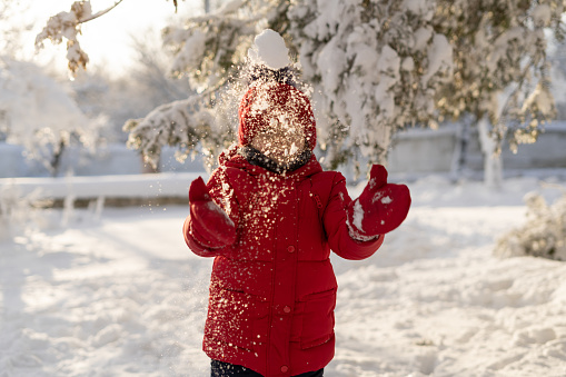 The girl plays with snow in winter. A little sister in a red jacket and knitted hat tears and catches snow in a winter park for Christmas. Children play in the snowy forest. Catching snowflakes