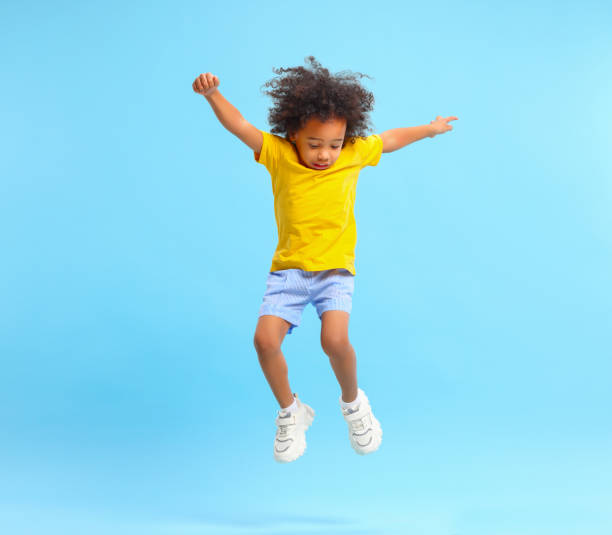 Active black kid jumping against blue background Full length of energetic little ethnic boy with Afro hairstyle in stylish casual outfit jumping with raised arms and looking down against blue background jumper stock pictures, royalty-free photos & images
