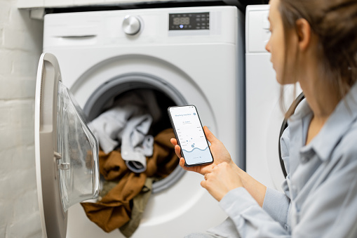Woman controls washing machine with a smartphone, holding phone with a running program for smart home appliances