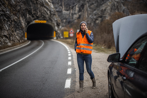 Young woman in reflective waistcoat standing next to car and calling roadside assistance