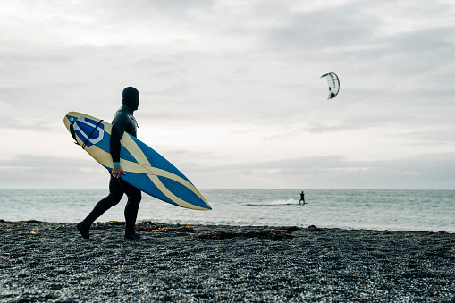 Man in wetsuit carrying surfboard and looking at sea. Surfer looking at sea while walking with surfboard on beach.