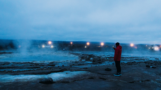 Panoramic view of a young man explorer in red jacket taking photo of the scenic volcanic area with geysers after the sun has gone down in South Iceland