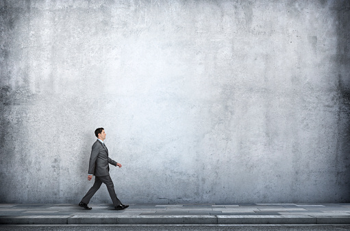 A businessman walks on a sidewalk next to a large concrete wall that provides ample room for copy and text.