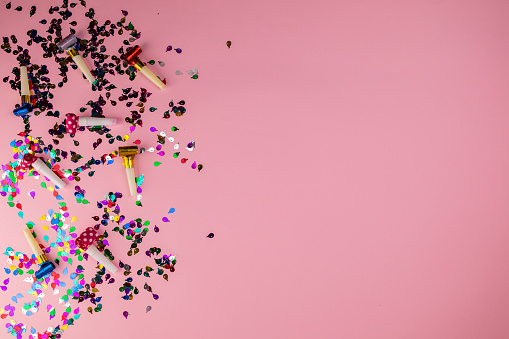 Multi-colored confetti and party horn blowers on pink background. Flat lay concept.