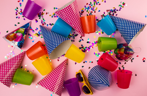 Multi-colored confetti, party hats and disposable cups on pink background. Flat lay concept.