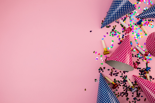 Multi-colored confetti, party hats and party horn blowers on pink background. Flat lay concept.