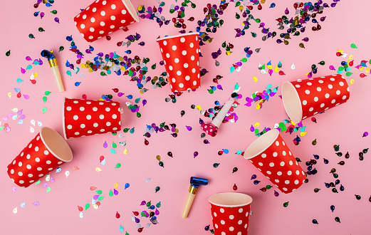 Multi-colored confetti, disposable cups and party horn blowers on pink background. Flat lay concept.