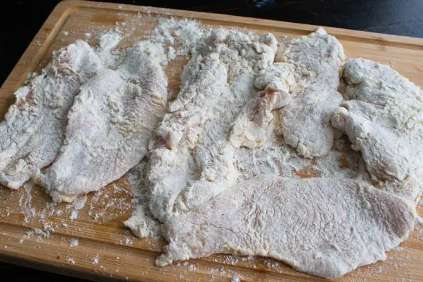 Raw thinly sliced chicken breasts covered in flour on a bamboo cutting board