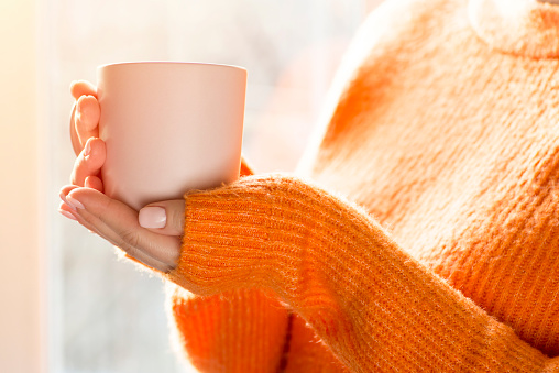Female hands with hot drink. A young woman in orange sweater holding cup of tea or coffee near window at home.