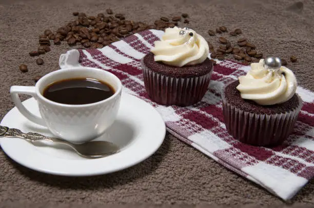 Two cupcakes on fabric background, white coffee cup, homemade cake with cream and roasted coffee beans. Selective focus.