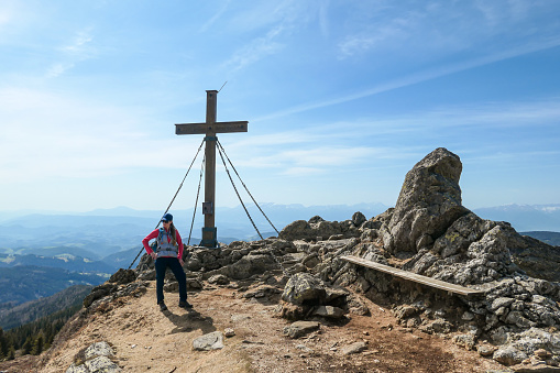 A woman with hiking backpack reaching the top of Sauofen in Austrian Alps. A wooden cross on top. Lots of stones around. Fall vibes. High mountain chains in the back. Serenity and achievement