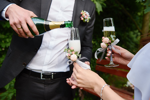 Groom in black suit pouring champagne from bottle into glass outdoors, copy space. Wedding couple in love