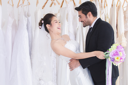 The Happiness couple newlywed beautiful Asean bride and mix race or foreigner groom hold hand togher dancing with fun in the wedding dress fitting room background, marriage with foreigner / wedding concept