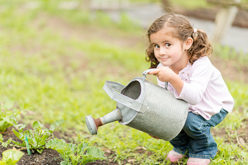 An adorable girl is watering the plants in her garden. She is using gardening gloves to hold the watering can. She is looking at the camera and smiling softly.