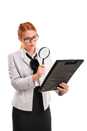 A portrait of a young businesswoman looking at a document on a clipboard through a magnifying glass, isolated on white background. Analysis, scrutiny concept.