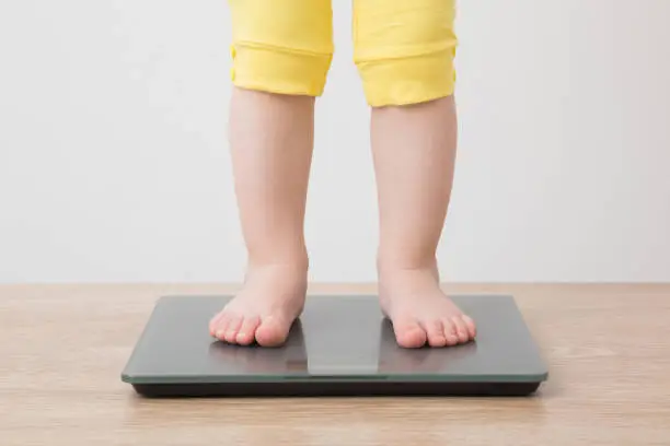 Photo of Baby girl with barefoot standing on weight scales on wooden floor at light gray wall background. Closeup. Front view. Care about body. Weight control concept.