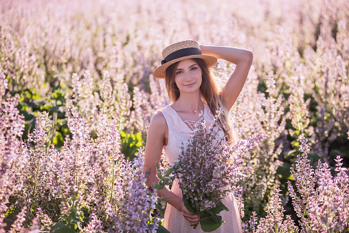 Young blonde woman in straw hat on the background of blooming field of pink sage. Close-up portrait beautiful girl holding bouquet flowers in hands. Weekend walk outside the city. Agricultural texture